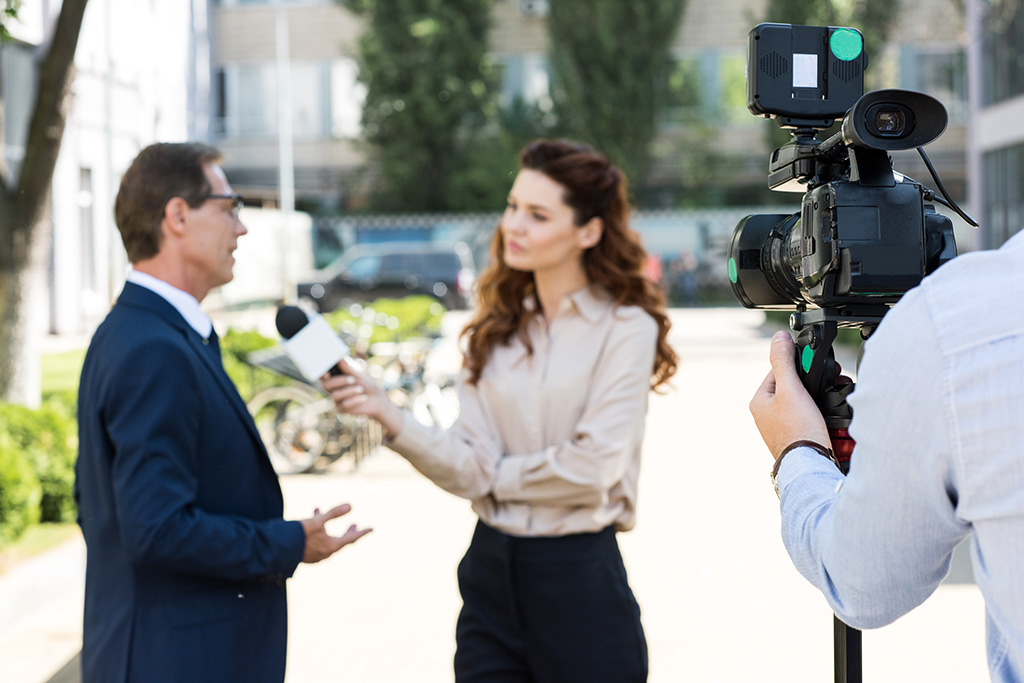 une journaliste femme qui tient un micro et qui interview un homme en costume, ils sont filmés par un caméraman