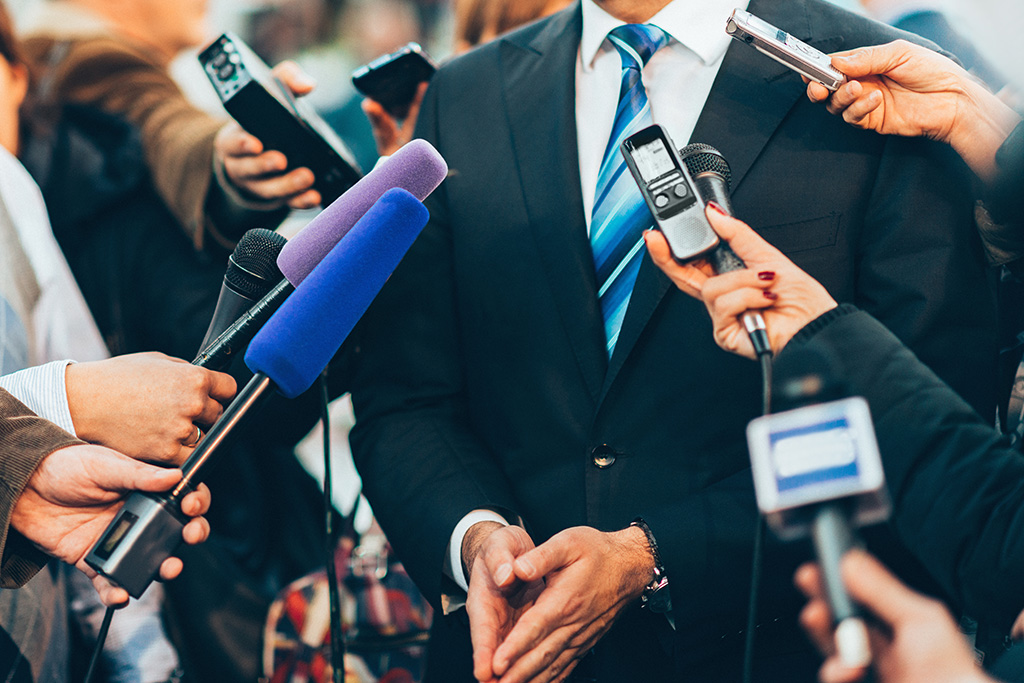 homme en costume dont on ne voit pas le visage et qui se fait interviewer par plusieurs journaliste dont on ne voit que les mains qui tiennent des micros ou des téléphones portables