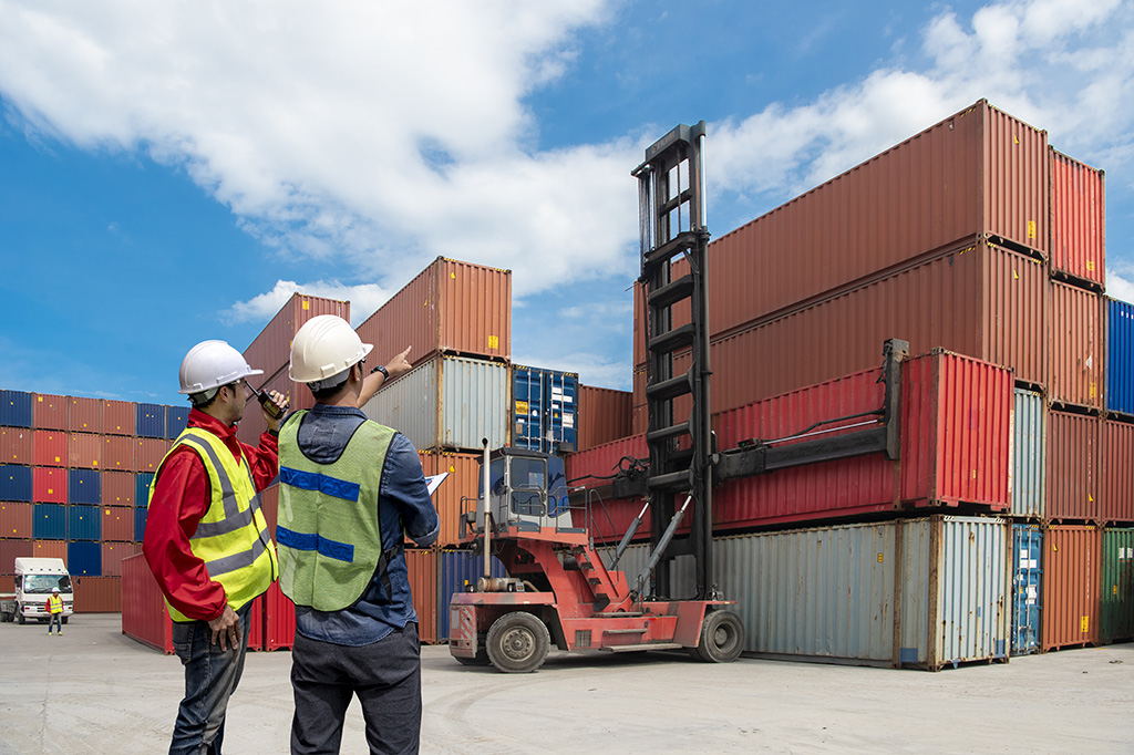 deux hommes portants un casque et un gilet jaune de sécurité responsable logistique sur un quai de chargement de containers regardant un véhicule chargeant des containers