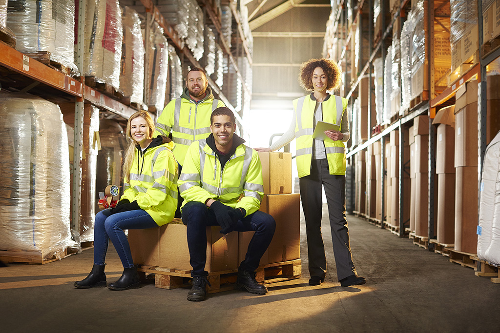 portrait dans un entrepôt d'une équipe logistique de 4 personnes dont deux hommes et deux femmes portant chacun un gilet jaune de sécurité et qui sourient tous