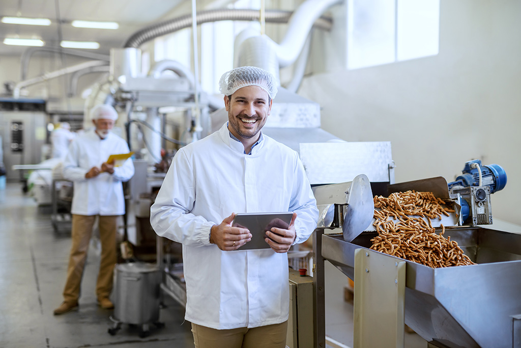 dans un entrepôt agro alimentaire homme souriant portant une blouse blanche et une charlotte tenant à la main une tablette numérique il est devant un chaine de production de produits alimentaires type pâtes 