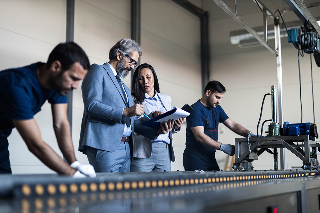 un homme en costume et une femme qui inspectent la ligne de production et deux hommes qui sont entrain de travailler sur cette ligne de production