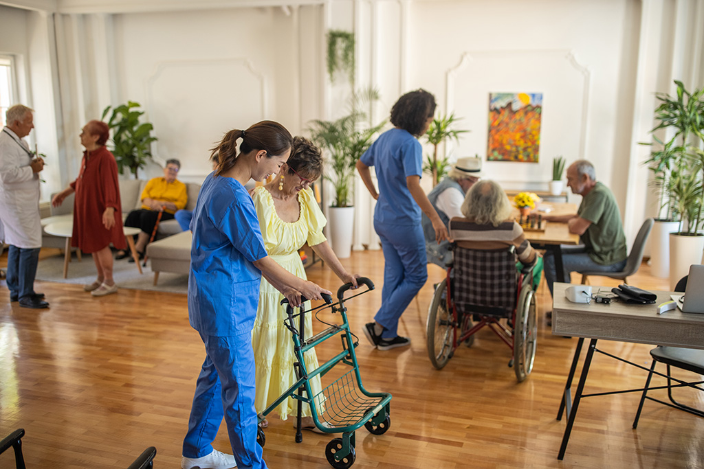 dans une salle de maison de retraite femme soignante aidant une femme âgée à marcher avec un déambulateur en arrière plan une femme soignante avec un groupe de 3 personnes âgées assises à une table