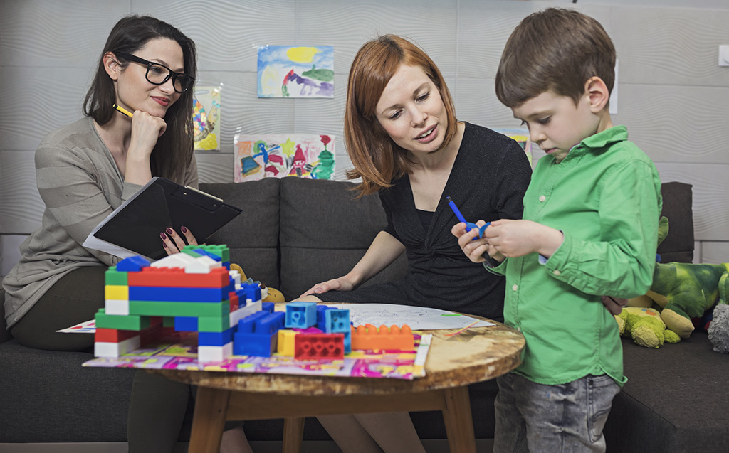 dans un bureau éducatrice jeunes enfants qui tient un bloc note et qui regarde en souriant un enfant qui joue accompagné par sa mère