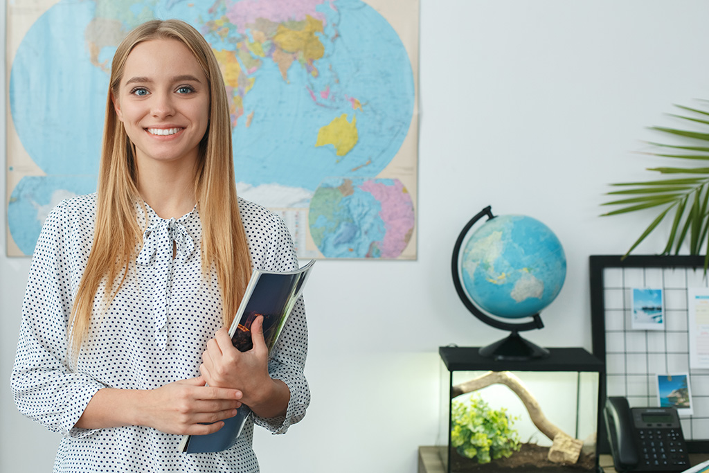 femme agent de voyage qui sourit debout devant une carte du monde et tenant un bloc note à la main en arrière plan avec un globe terrestre un planning et un téléphone posés sur un bureau