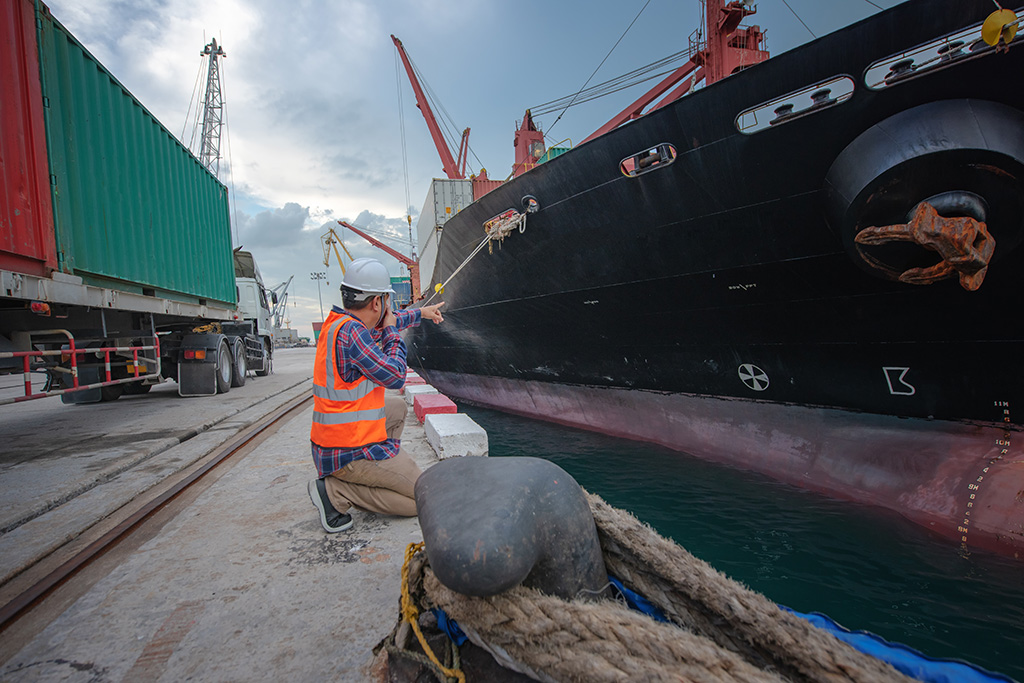 agent de transport fluvial portant un casque de chantier un gilet de sécurité orange parlant au talkie walkie et en arrière plan un navire de marchandise et un porte container