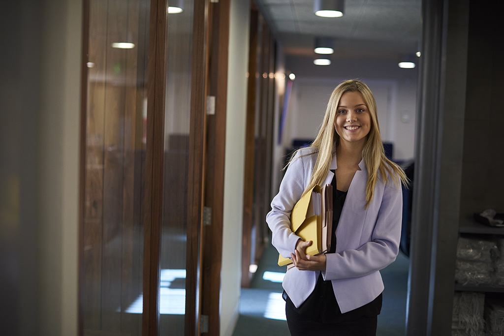 secrétaire souriante en tailleur debout dans un couloir desservant des bureaux. Elle tient plusieurs dossiers sous le bras