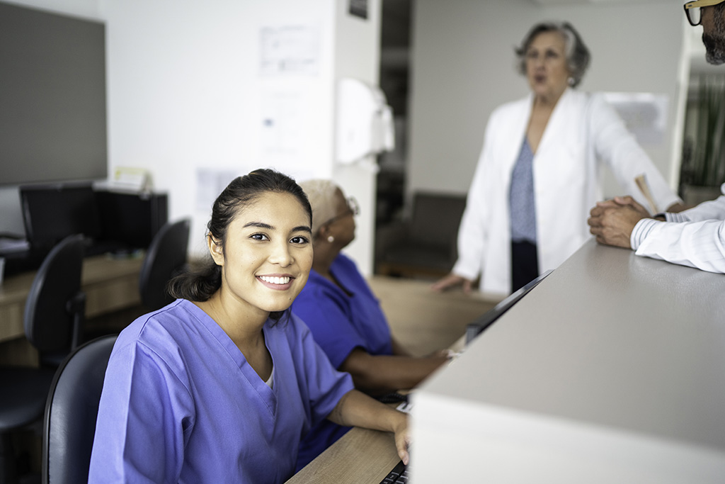 secrétaire médicale en blouse bleu souriante assise derrière son bureau et en arrière plan le médecin en blouse blanche