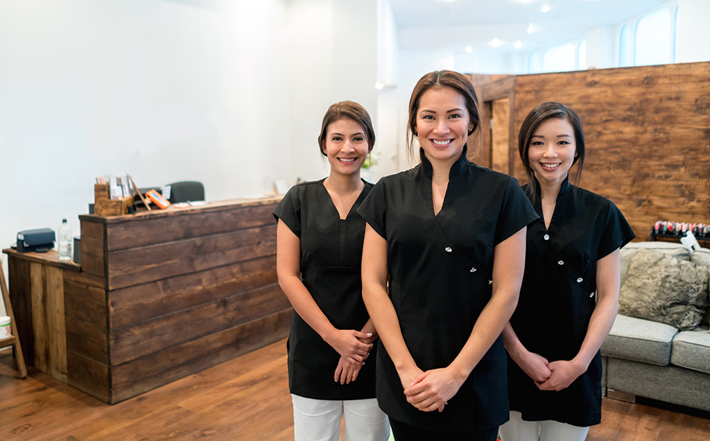 équipe de trois esthéticiennes souriantes en blouse noire se tenant à l'entrée de l'institut de beauté