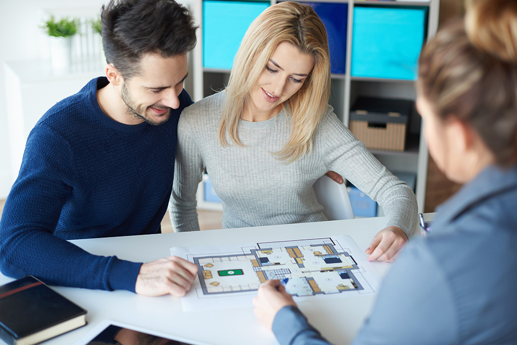 dans une agence immobilière, un couple et un agent immobilier femme touts assis à un bureau et regardant ensemble les plans intérieurs d