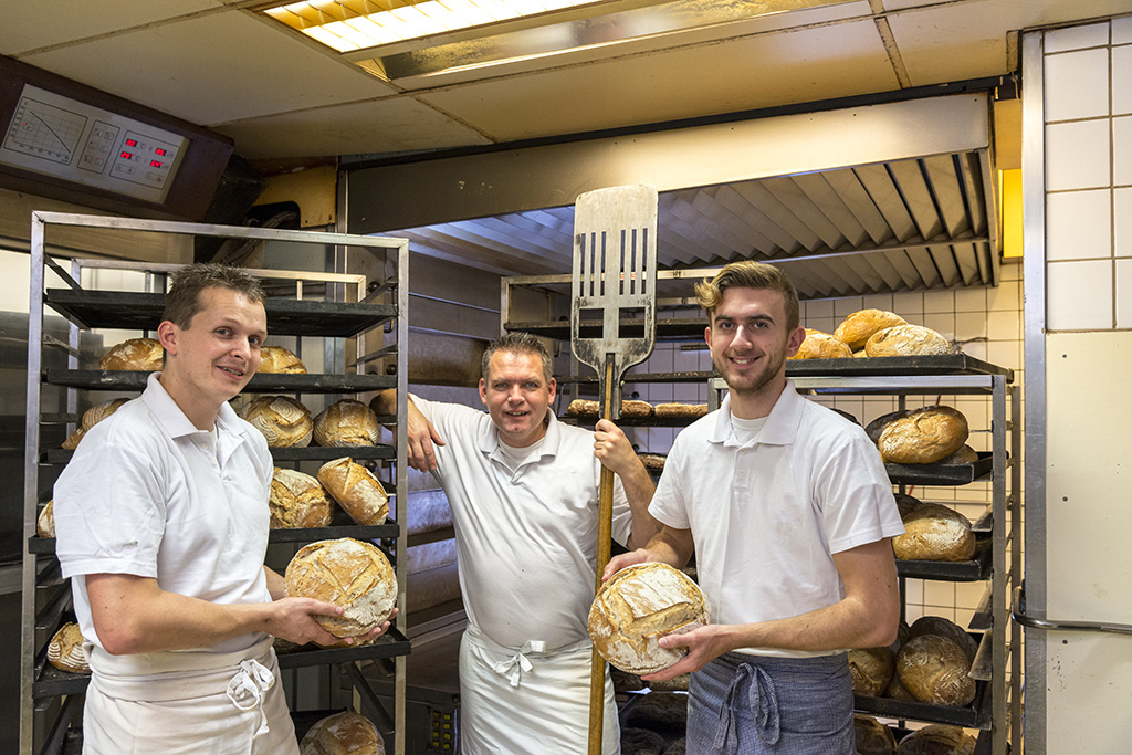 trois boulangers tenant des pains en main dans leur boulangerie en arrière plan des chariots de pains