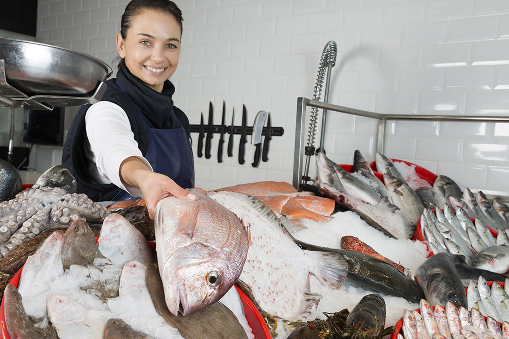 poissonnière en habit tablier souriante derrière son présentoir à poisson et en tenant un des poissons en main