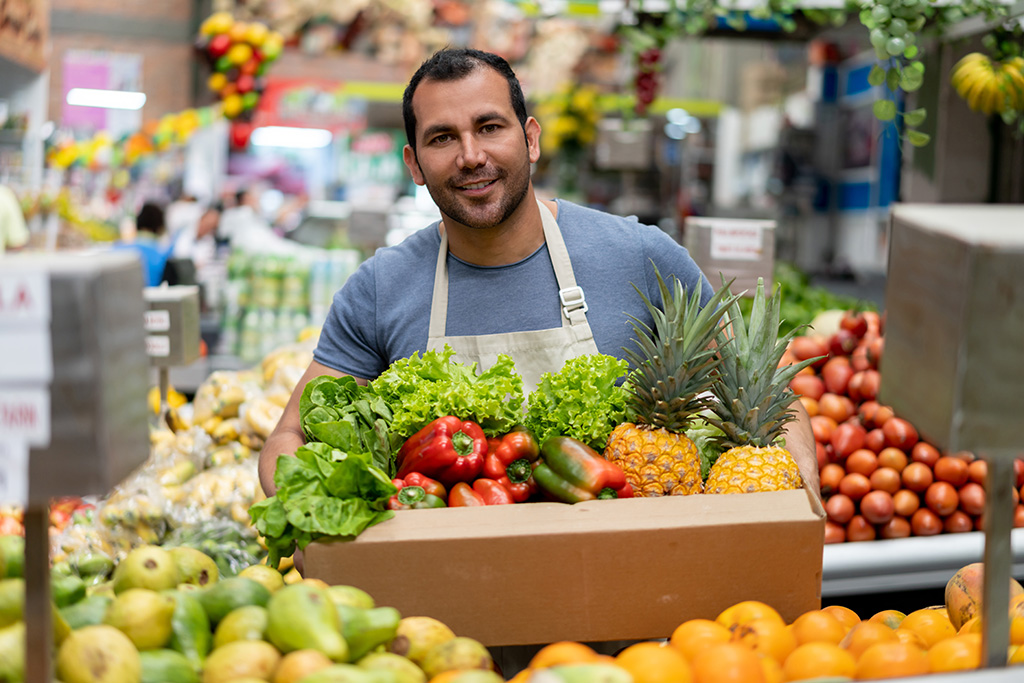 primeur derrière son stand de fruits et légumes sur un marché qui sourit et qui tient dans les mains une cagette de légumes 