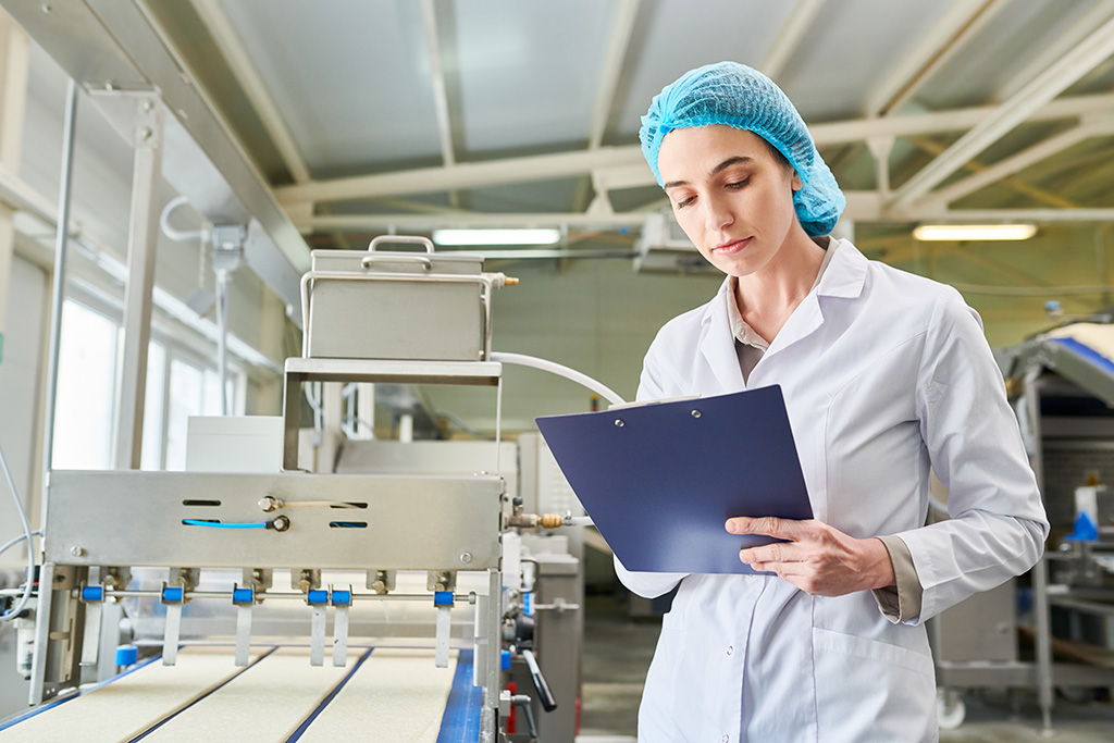 Portrait d’un technologue femme portant une blouse blanche et une charlotte tenant un porte document vérifiant la machine de fabrication de la production de pâte